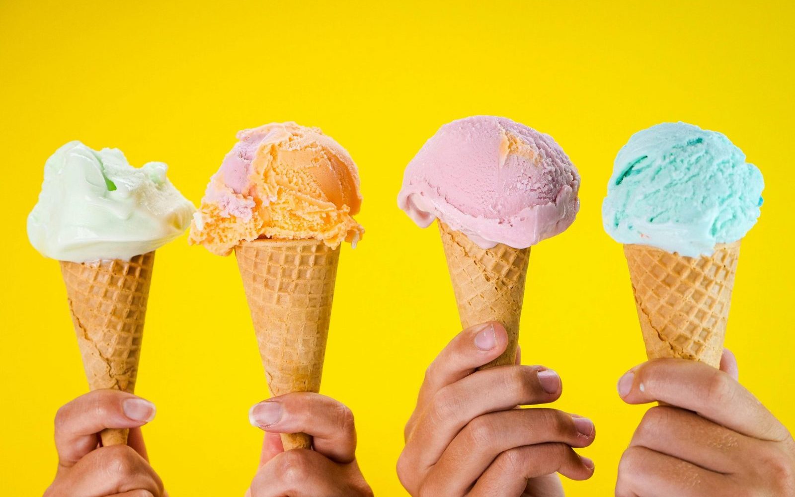 Four colorful ice cream cones held up against a bright yellow backdrop, showcasing various flavors.